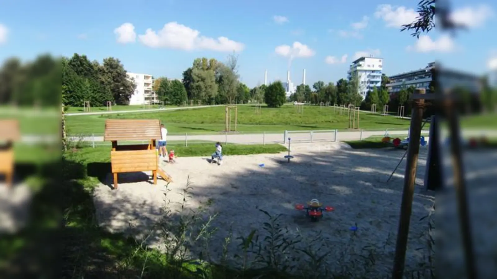 Der zum Schutz der Kinder vor Radlern und Hunden eingezäunte Spielplatz im Zentrum der Parkanlage im Bogenhauser Ortsteil Johanneskirchen. Im Hintergrund die Schlote des Heizkraftwerks München Nord. 	 (Foto: hgb)