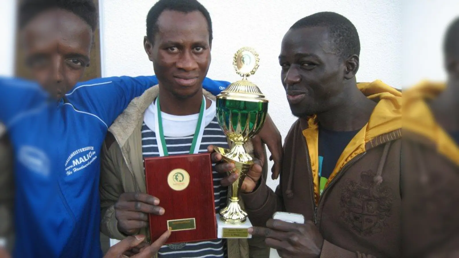 Trainer Tangara Boubacar (Mitte), Mannschaftskapitän Diaby Abdou Khadry (r.) und Spieler Sadio Mane (l.) zeigen die Pokale aus den buntkicktgut-Turnieren. (Foto: Helferkreis_Mainaustraße)