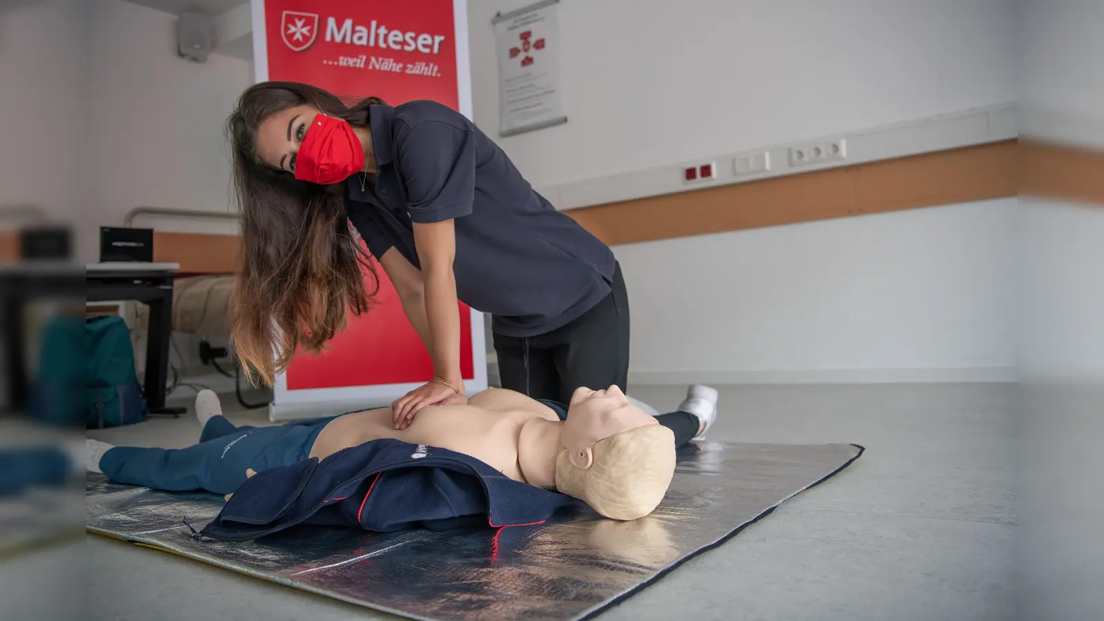 Eine Erste Hilfe Ausbilderin der Malteser macht die Herz-Lungen-Wiederbelebung vor. Aus hygienischen Gründen trägt sie eine Mund-Nasen-Bedeckung. (Foto: Malteser/Gerhard Seeger)