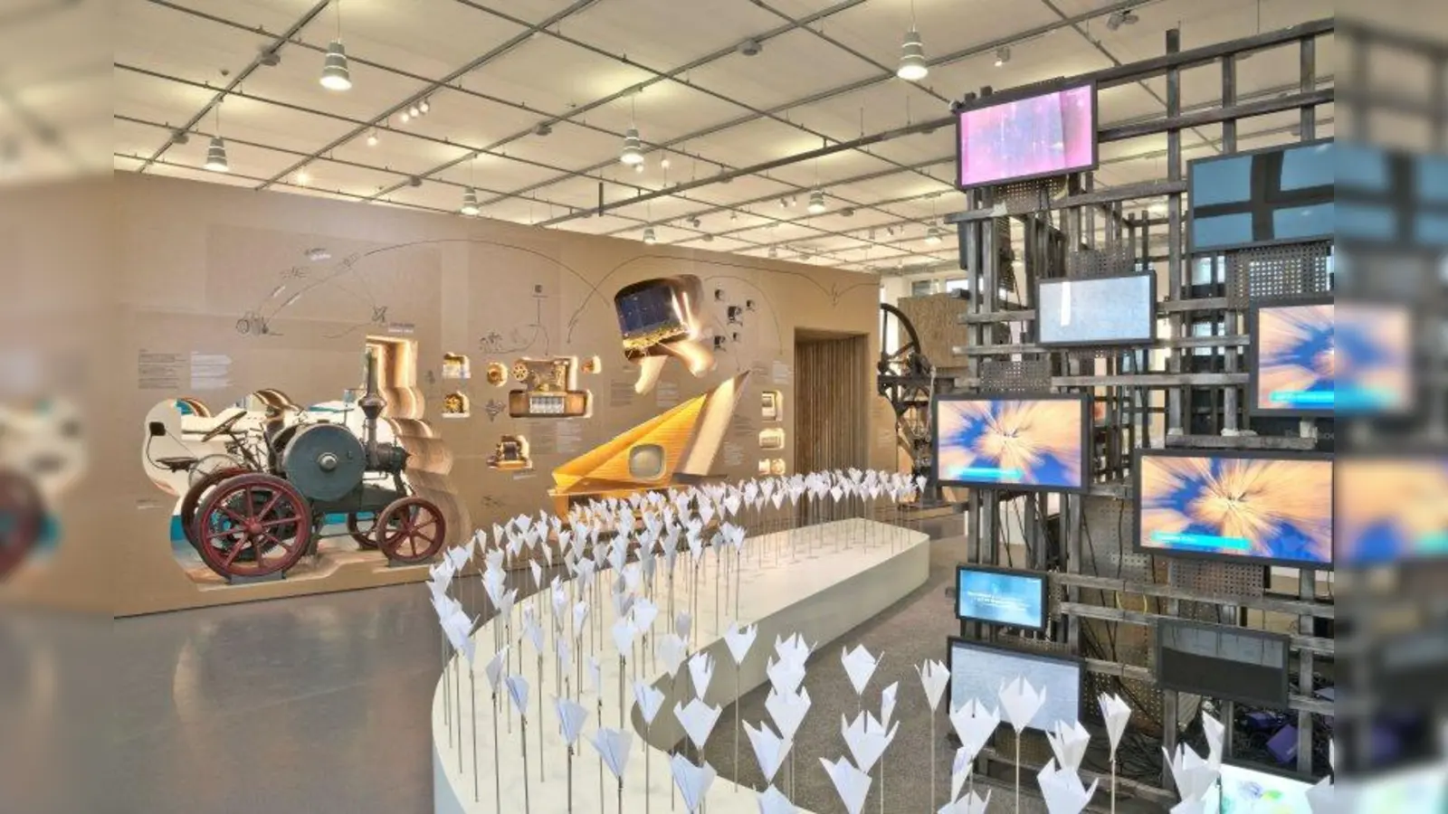 Das große Auftaktelement einer künstlichen Landschaft, in der fast 1000 Papierblumen blühen, lädt mit Sitzmöglichkeiten zum Verweilen ein. Im Blickfeld ist ein großer Medienwürfel in Form eines Stahlgerüsts. Auf 61 Monitoren führen Filme in die Themen des Anthropozäns ein. (Foto: Deutsches Museum)