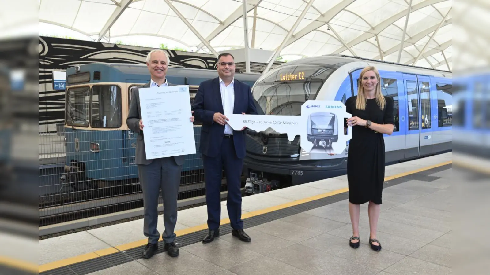 Konrad Schober (l.) mit der Inbetriebnahme-Genehmigung neben Ingo Wortmann, dem Deborah Schmitz (Siemens Mobility) einen symbolischen U-Bahn-Schlüssel überreicht. Sie präsentieren sich vor dem 85. C2-Zug und einem Zug der Serie A, die vom den C2-Zügen ersetzt wird. (Foto: SWM/ MVG)