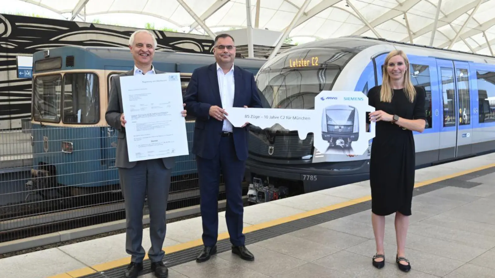 Konrad Schober (l.) mit der Inbetriebnahme-Genehmigung neben Ingo Wortmann, dem Deborah Schmitz (Siemens Mobility) einen symbolischen U-Bahn-Schlüssel überreicht. Sie präsentieren sich vor dem 85. C2-Zug und einem Zug der Serie A, die vom den C2-Zügen ersetzt wird. (Foto: SWM/ MVG)