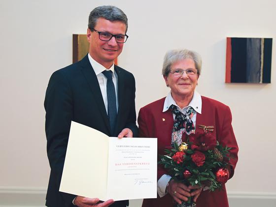 Staatssekretär Bernd Sibler präsentiert die Urkunde, das Bundesverdienstkreuz heftet bereits am Revers von Roswitha Bendl.	Foto: © StMBW