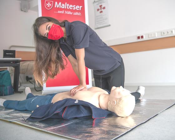 Eine Erste Hilfe Ausbilderin der Malteser macht die Herz-Lungen-Wiederbelebung vor. Aus hygienischen Gründen trägt sie eine Mund-Nasen-Bedeckung. Foto: Malteser/Gerhard Seeger